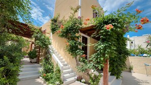 Un edifici amb façana color beix, escales blanques cap a una porta de fusta, i un jardí amb plantes i flors variades.
