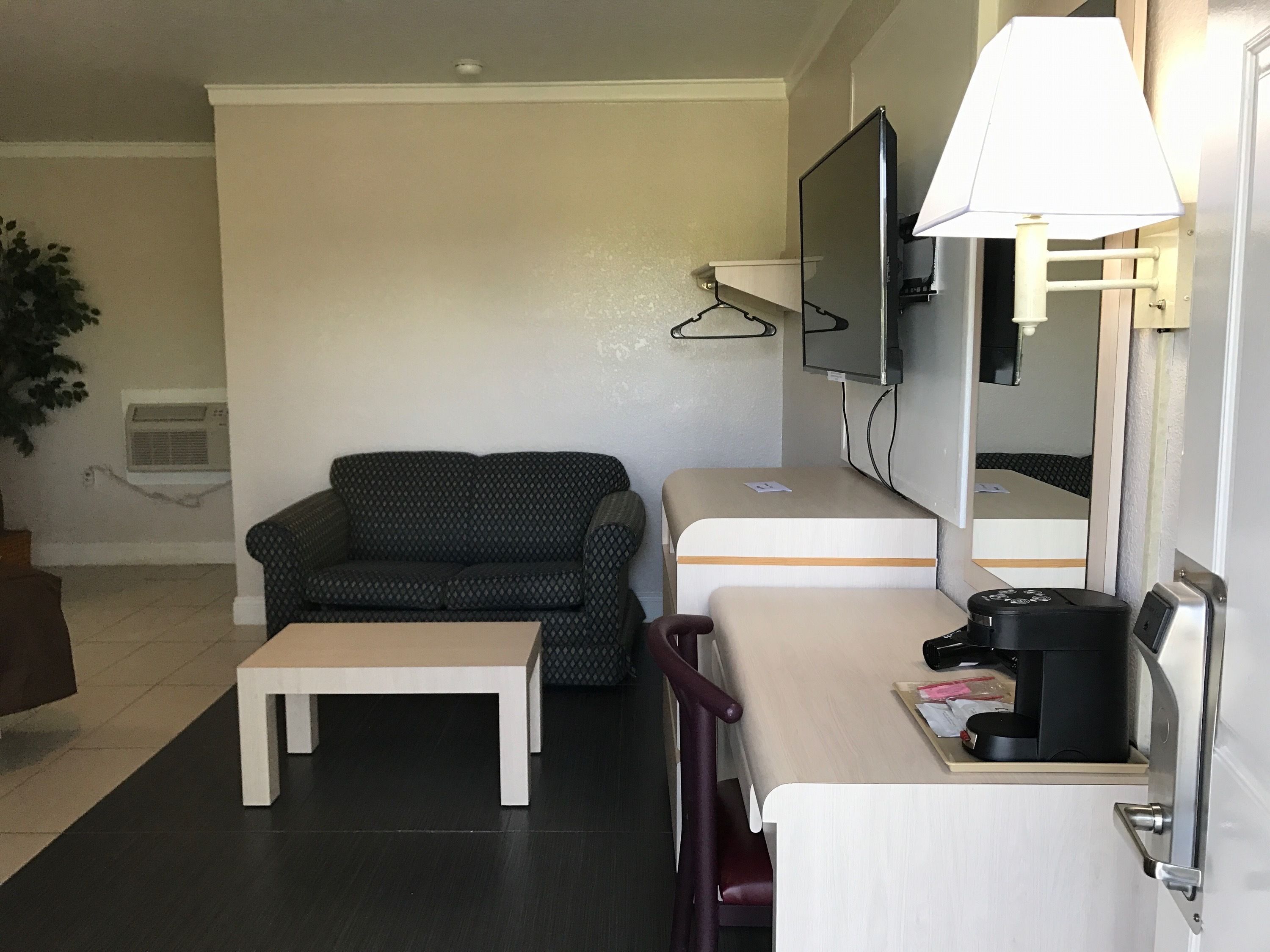 Ein kleines Hotelzimmer mit einer Sitzecke, einem Tisch, einem Schreibtisch mit Lampe, einem Fernseher und einem Spiegel.