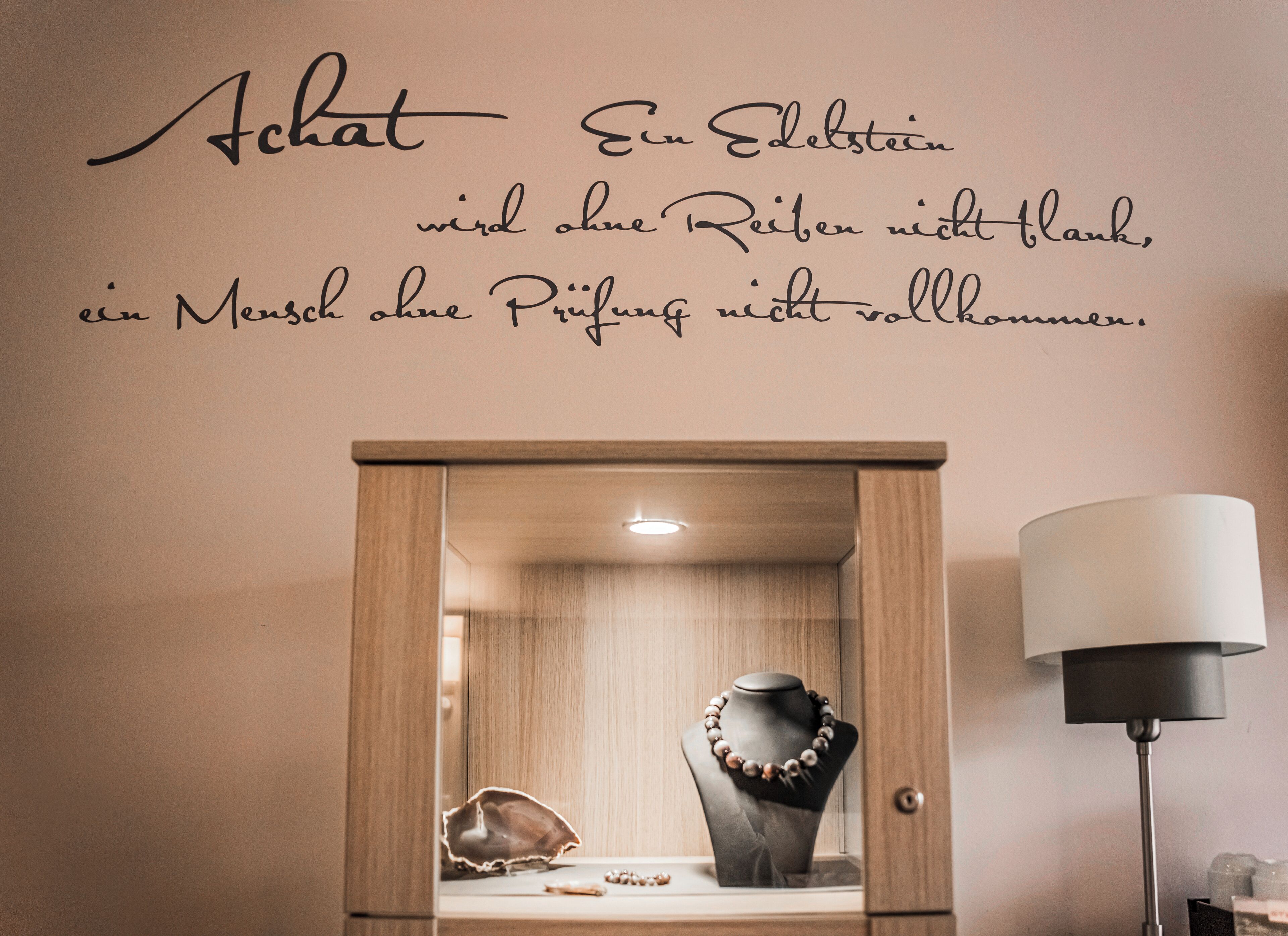 Eine Wand mit einem Zitat in Deutsch, eine Holzkassette mit Schmuck und eine Stehlampe.