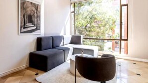 Un salon moderne avec un canapé d’angle, une petite table blanche et un fauteuil rond noir. Une grande fenêtre offre une vue sur la verdure à l’extérieur.