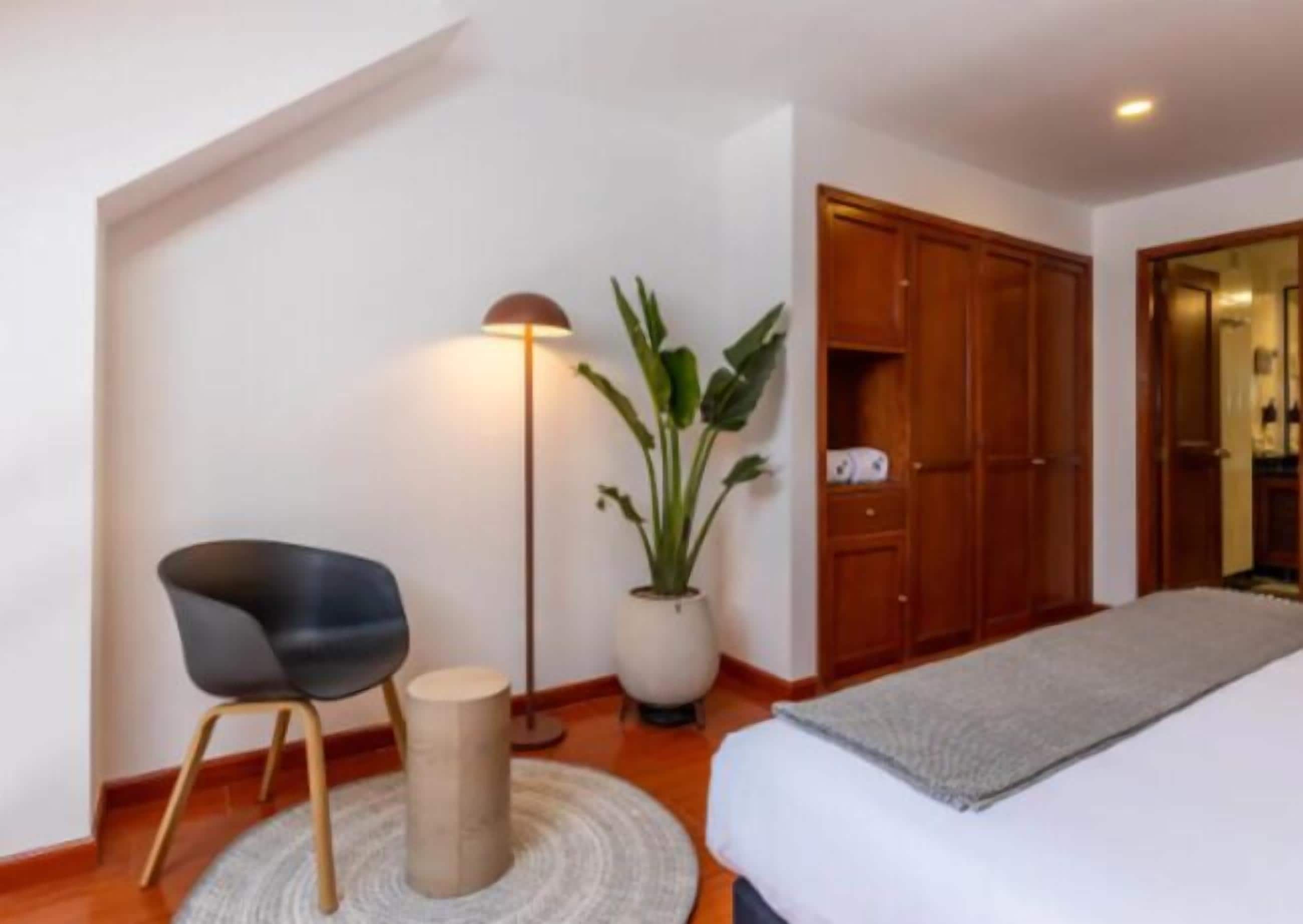 Un dormitorio con cama, escritorio, silla, lámpara de pie, planta en maceta y armario.