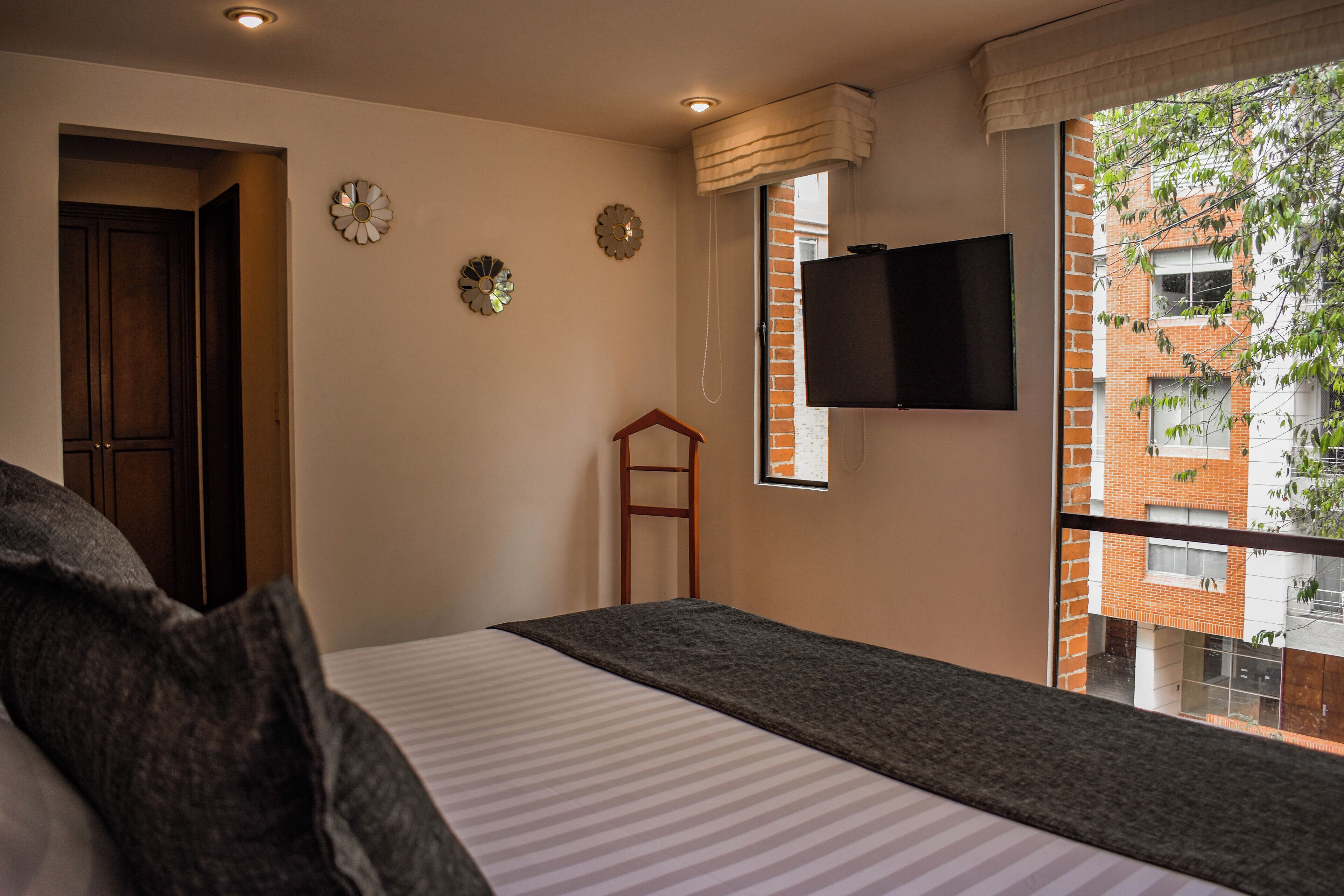 Une chambre à coucher avec un lit, un téléviseur fixé au mur et une fenêtre donnant sur des immeubles.