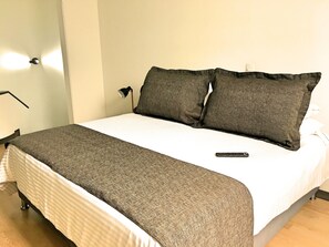 Un lit bien fait, recouvert d’une couverture à rayures blanches et brunes, avec deux oreillers foncés et une télécommande posée sur le lit.