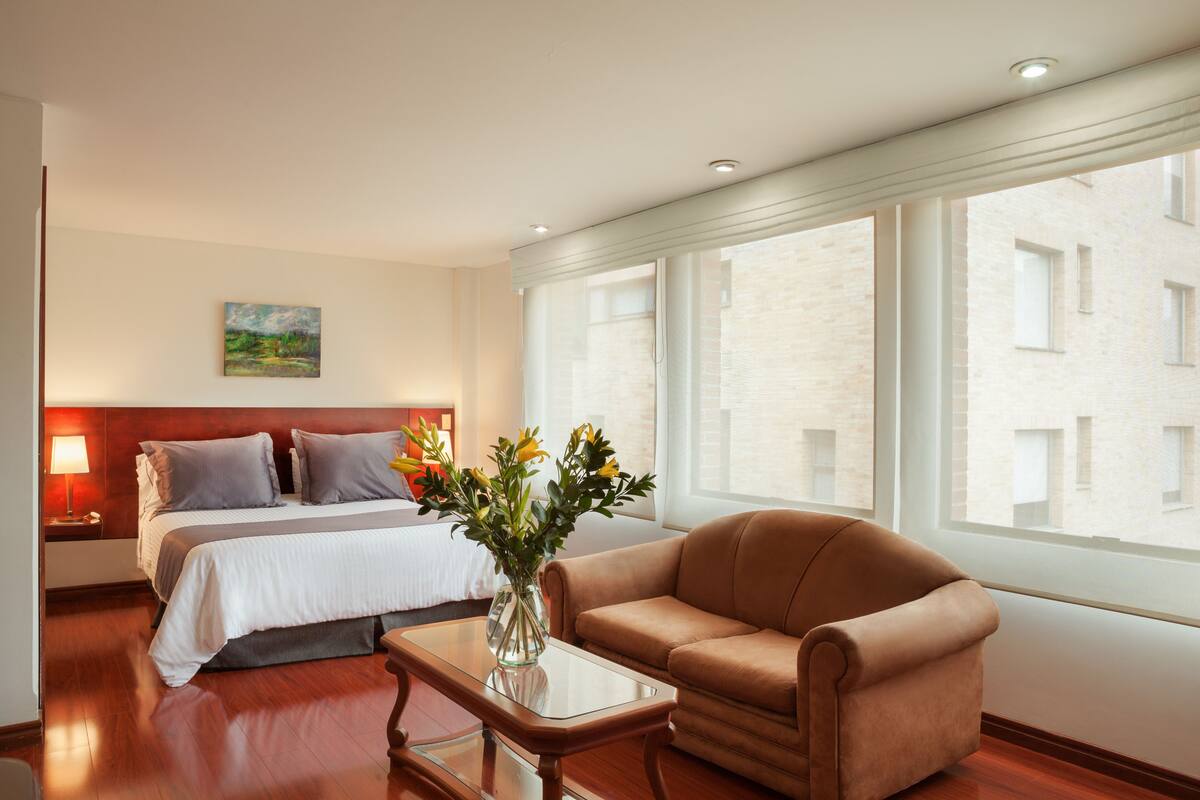 Una camera d'albergo con un letto, un divano, un comodino con una lampada, un vaso con fiori e ampie finestre che offrono vista sugli edifici.