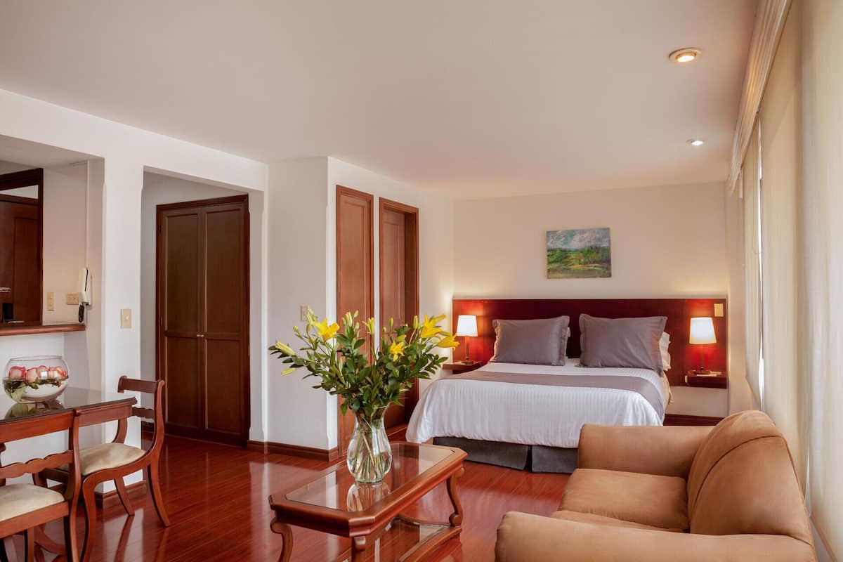 Ein Hotelzimmer mit einem Bett, Nachttischlampen, einem Schreibtisch mit Blumenvase, einem Sessel und einem Bild an der Wand.