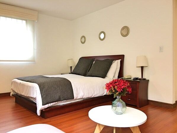 Une chambre d’hôtel bien rangée, avec un lit, des lampes de chevet, une petite table sur laquelle est posé un vase, et une fenêtre avec des stores.