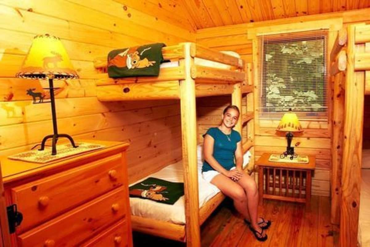 Une chambre rustique en bois avec un lit superposé, une table de chevet, une lampe et une fenêtre avec des stores.