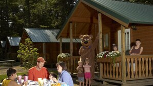 A wooden cabin with a porch, a table with food and drinks, and a mascot. - Yogi Bear's Jellystone Park Camp-Resort Wisconsin Dells (Wisconsin Dells)