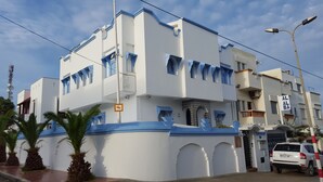 Un edificio de varios pisos con paredes blancas y detalles azules, que cuenta con una entrada protegida por una reja y un vehículo visible.