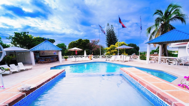 A swimming pool with lounge chairs and umbrellas, surrounded by palm trees and flags. - Visa Lodge (Port-au-Prince)