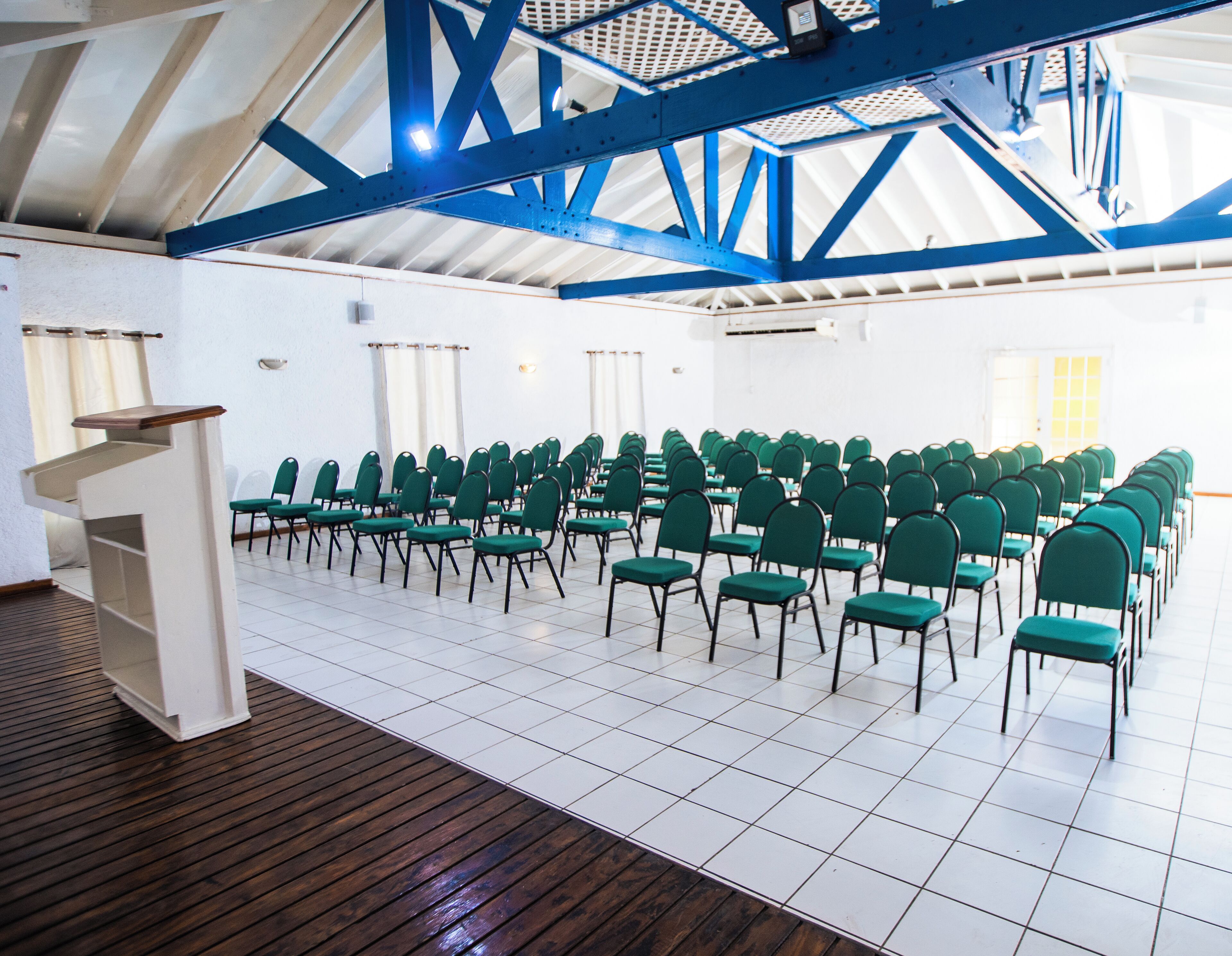A conference room with rows of green chairs, a podium, and a high ceiling with blue beams.
