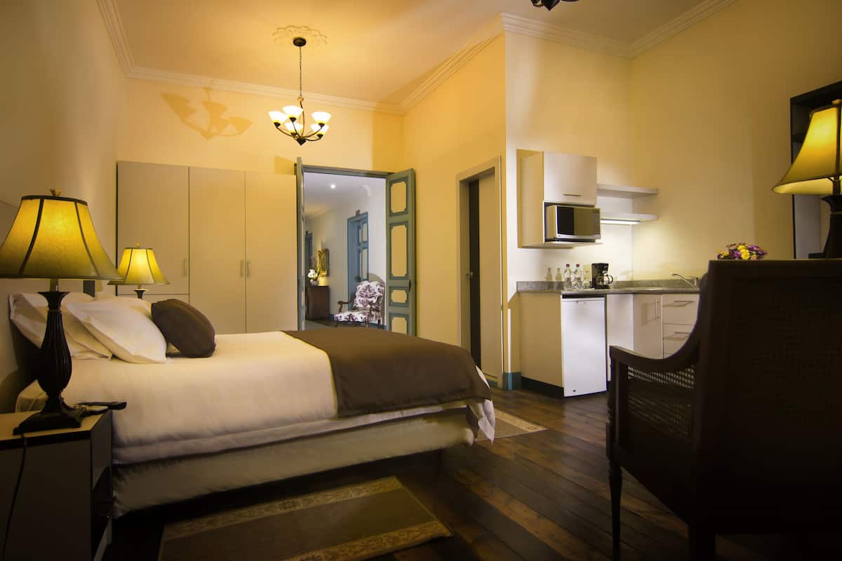 A hotel room with a bed, bedside lamps, a chair, a kitchen area with a microwave, and a door leading to another room.