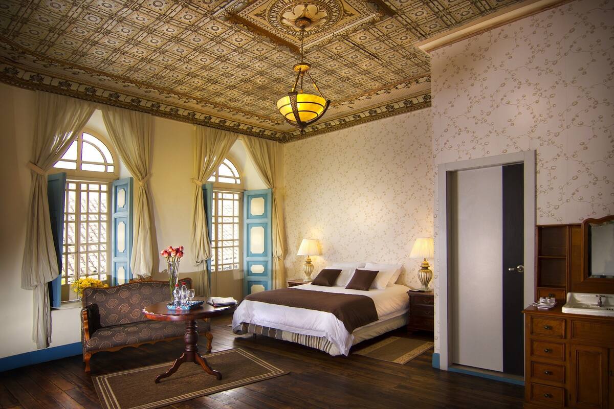 A room with a bed, a sofa, a chandelier, and a window with curtains.