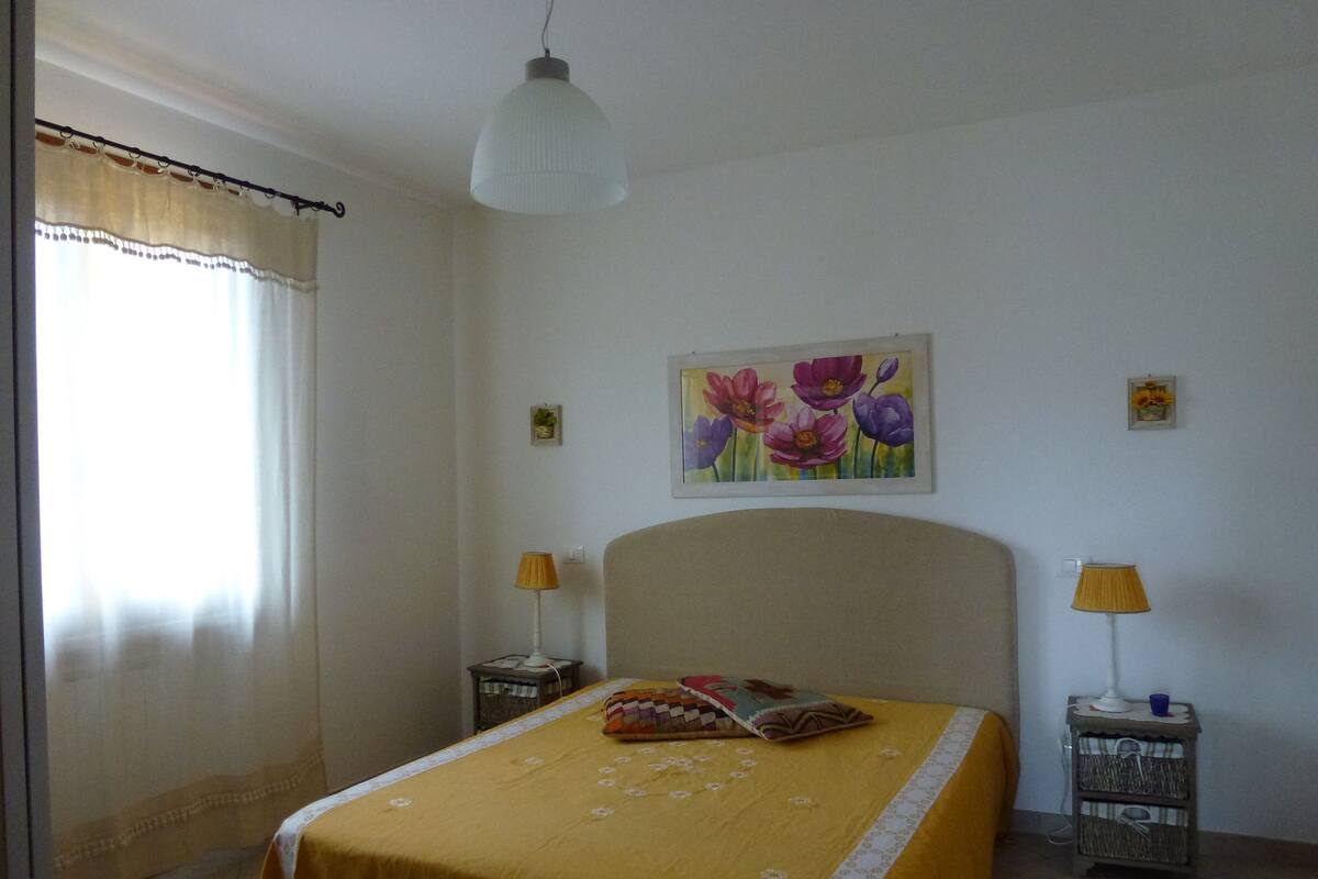 Une chambre à coucher avec un lit, deux tables de chevet, une lampe suspendue, un tableau et une fenêtre avec des rideaux.