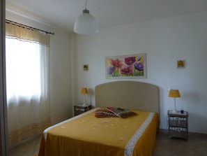 Une chambre à coucher avec un lit, deux tables de chevet, une lampe suspendue, un tableau et une fenêtre avec des rideaux.