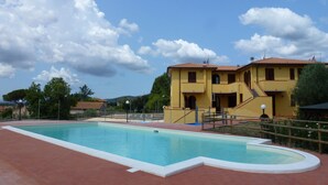 A swimming pool area with a yellow building in the background. - La Fonte degli Angeli (Suvereto)