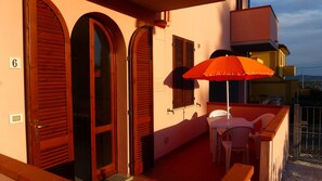Un balcon avec une table et des chaises, un parasol rouge, et un immeuble doté de fenêtres en arc de cercle et de volets.