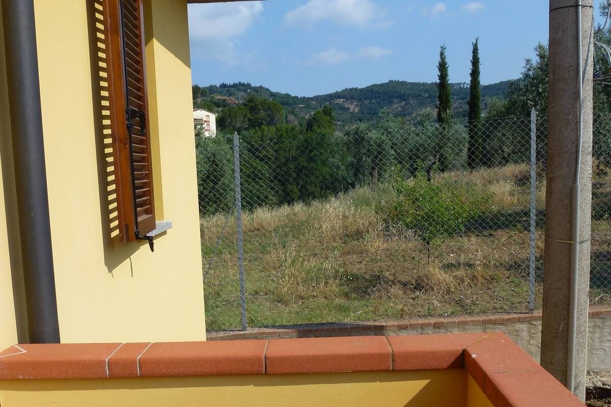 Un bâtiment jaune avec une fenêtre donnant sur un espace clos, agrémenté d’arbres et de collines.