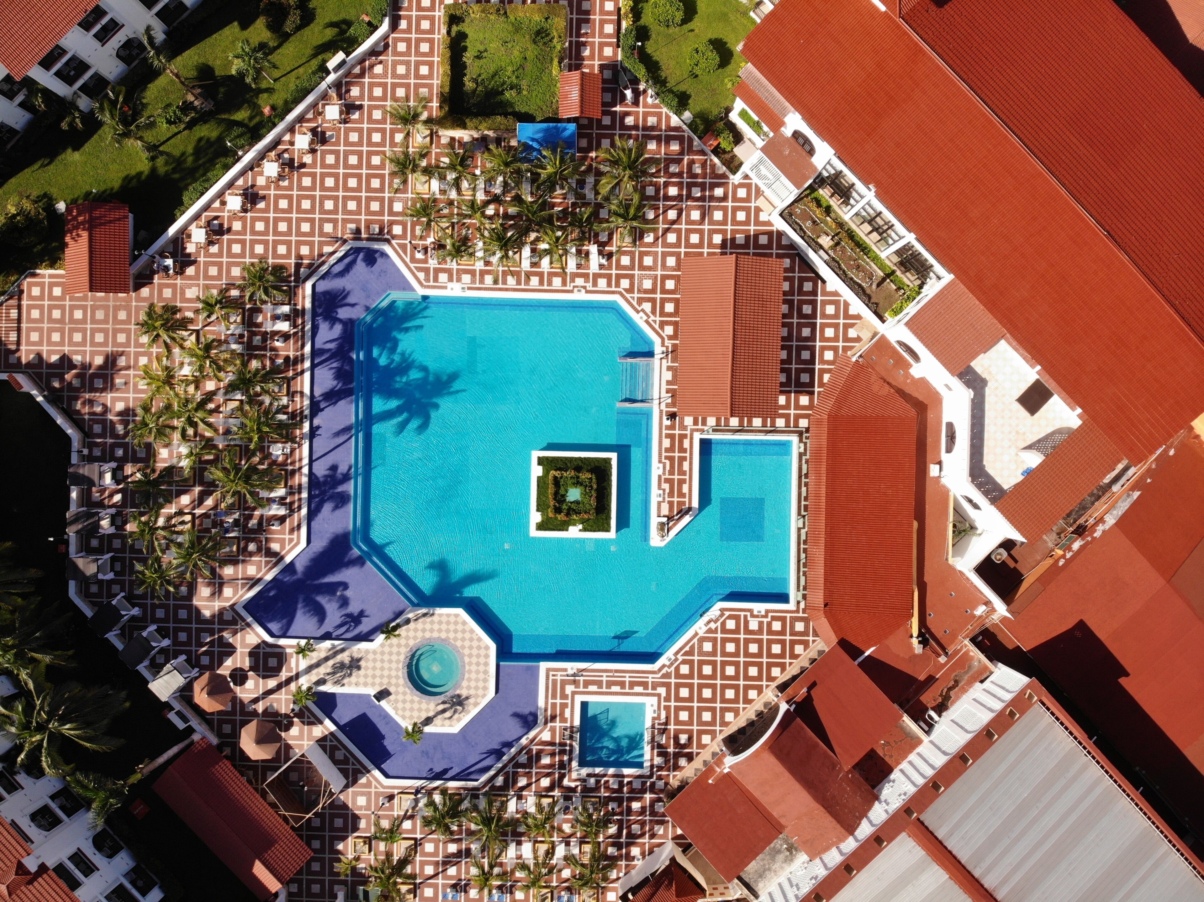 An aerial view of a resort with a central swimming pool, palm trees, and a patterned roof.