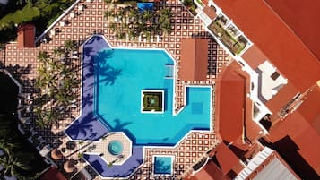 An aerial view of a resort with a central swimming pool, palm trees, and a patterned roof.
