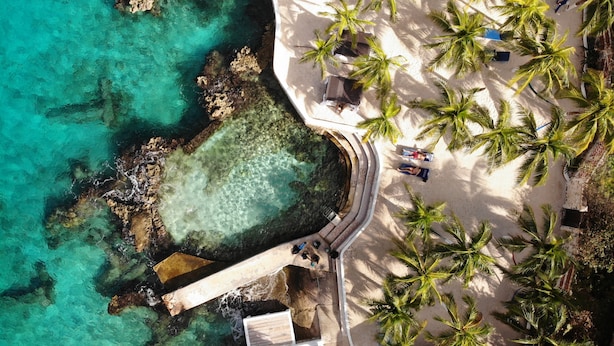 Vista aérea de una playa con agua turquesa cristalina, palmeras y un sendero que lleva a un edificio.