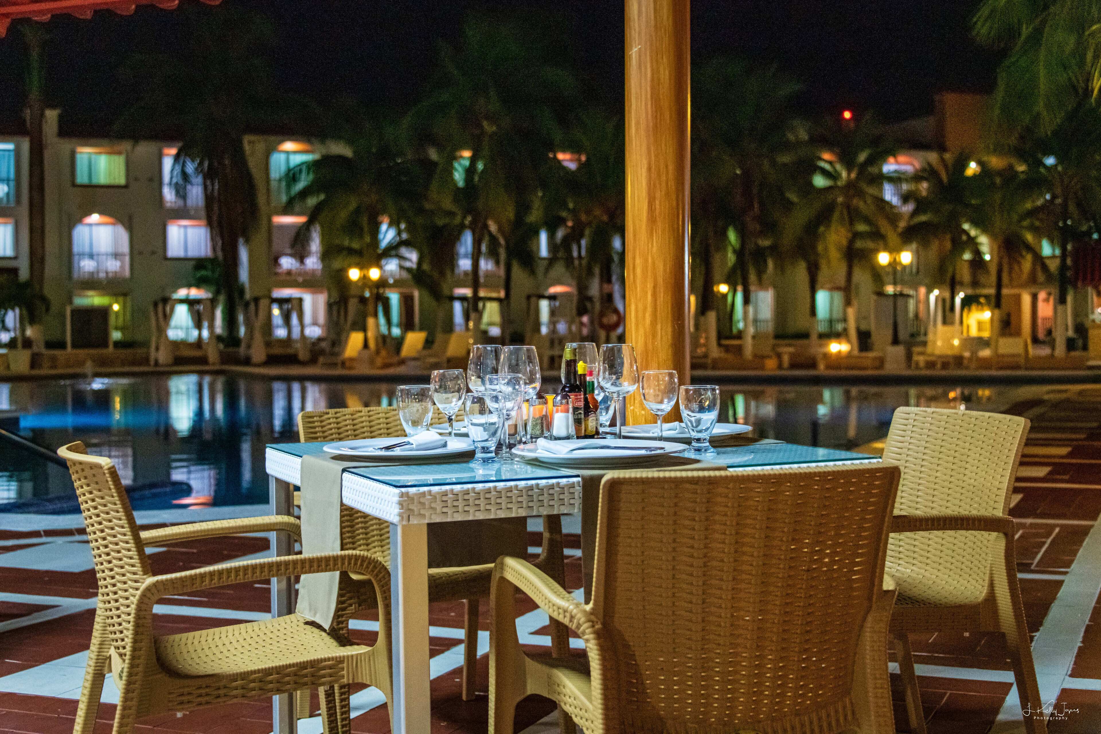 A dining table set with chairs and glasses, overlooking a pool with palm trees and buildings in the background.