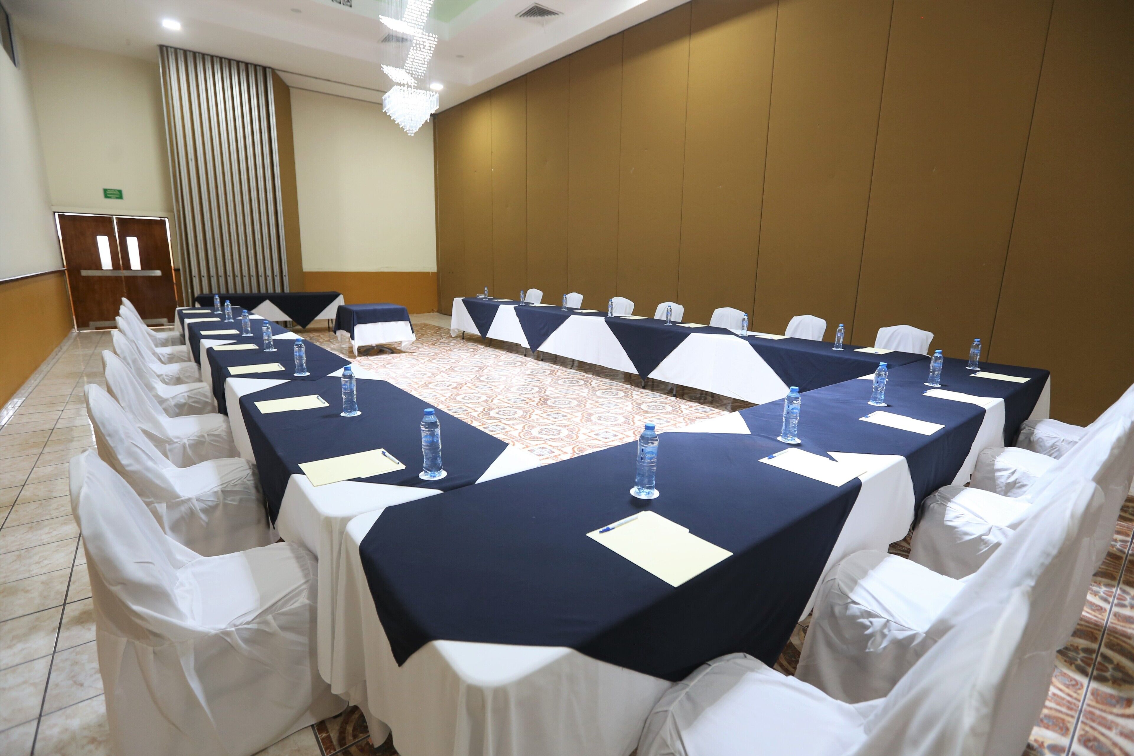 Uma sala de conferências com mesas compridas, cadeiras brancas e garrafas de água.