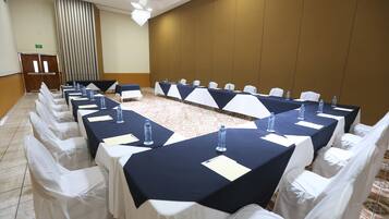 Uma sala de conferências com mesas compridas, cadeiras brancas e garrafas de água.
