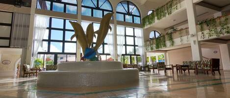 A spacious lobby with a central fountain, large arched windows, and a second-floor balcony with plants.
