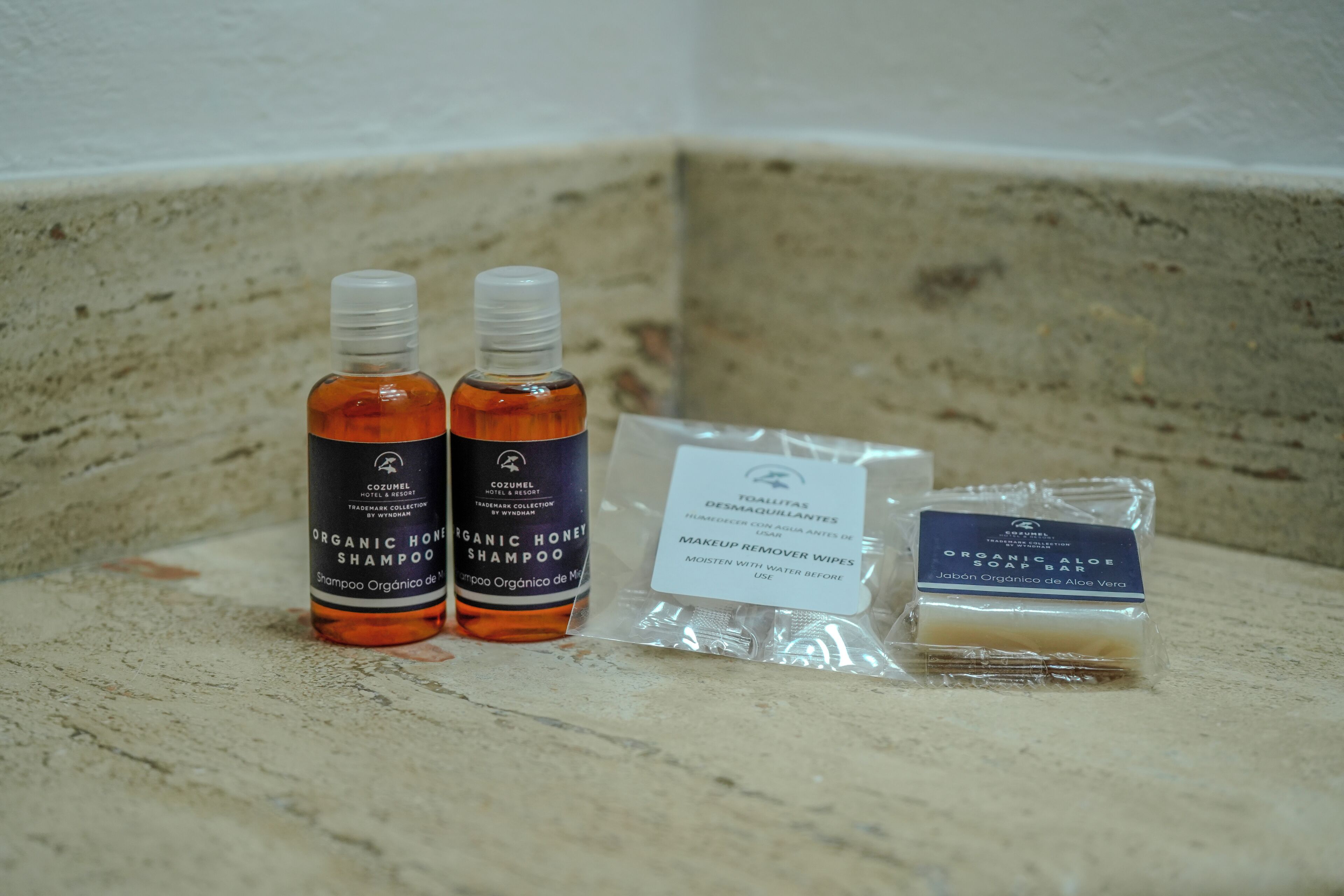 A bathroom counter with two bottles of organic honey shampoo, a package of makeup remover wipes, and a bar of organic aloe soap.