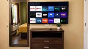 A hotel room with a large flat-screen TV displaying various streaming apps and channels, a wooden dresser with drawers, a potted plant, and a window with curtains. - Texas Bungalows Hotel & Hotel Que at Lakeline Austin Cedar Park (Cedar Park)