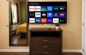 A hotel room with a large flat-screen TV displaying various streaming apps and channels, a wooden dresser with drawers, a potted plant, and a window with curtains. - Texas Bungalows Hotel & Hotel Que at Lakeline Austin Cedar Park (Cedar Park)