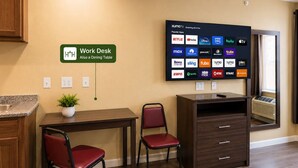 A work desk with a sign indicating it can also be used as a dining table, a flat-screen TV displaying various streaming apps, and a potted plant on the desk. - Texas Bungalows Hotel & Hotel Que at Lakeline Austin Cedar Park (Cedar Park)
