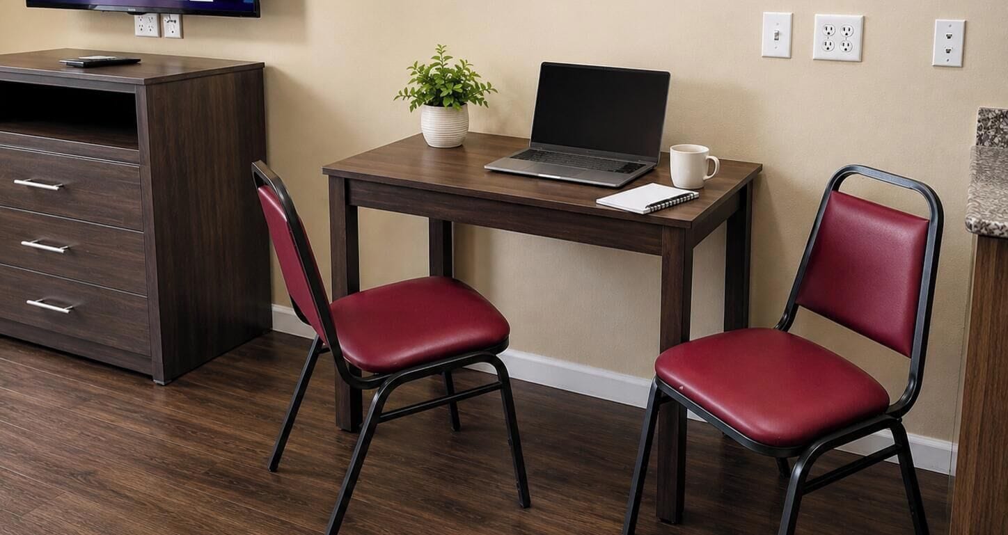 Un petit bureau comprenant un bureau, deux chaises rouges, un ordinateur portable, une plante en pot, un carnet et une tasse à café.