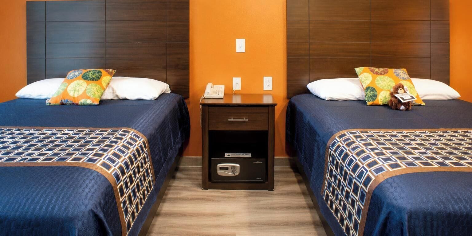 A hotel room with two beds, each with patterned bedding and pillows, a nightstand with a phone and a digital device, and a wall with a single light switch.