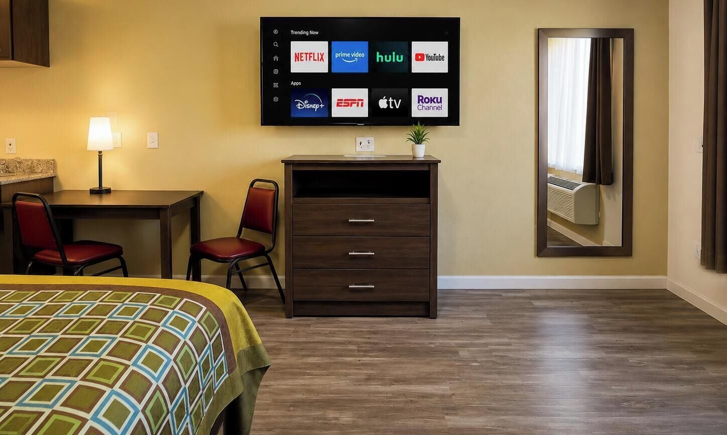 Habitación de hotel con cama, escritorio con lámpara, televisor mostrando servicios de streaming, cómoda y espejo.