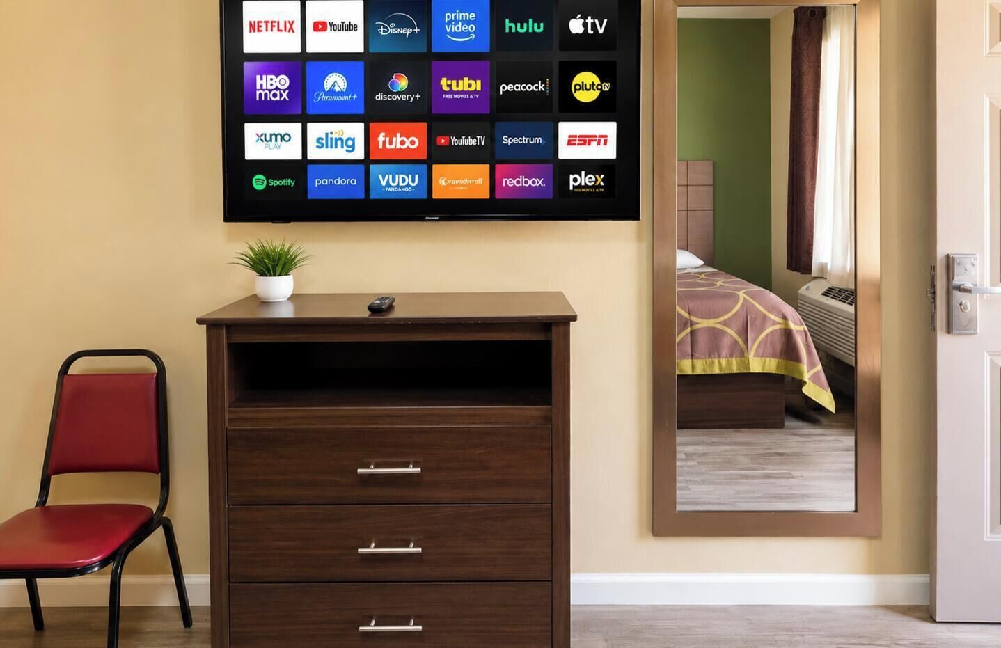 Ein Hotelzimmer mit einem großen Flachbildfernseher, der Logos verschiedener Streaming-Dienste anzeigt, einer Holzkommode mit Schubladen, einem roten Sessel und einer Topfpflanze.