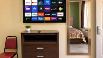 Ein Hotelzimmer mit einem großen Flachbildfernseher, der Logos verschiedener Streaming-Dienste anzeigt, einer Holzkommode mit Schubladen, einem roten Sessel und einer Topfpflanze.