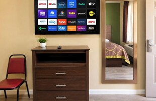 Quarto de hotel com uma televisão de tela plana grande exibindo logotipos de diversos serviços de streaming, uma cômoda de madeira com gavetas, uma cadeira vermelha e uma planta em vaso.