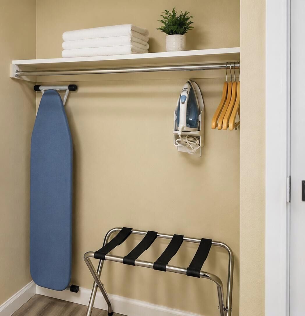 A room with a wall-mounted ironing board, a hanging rack with towels and a plant, and a metal drying rack.