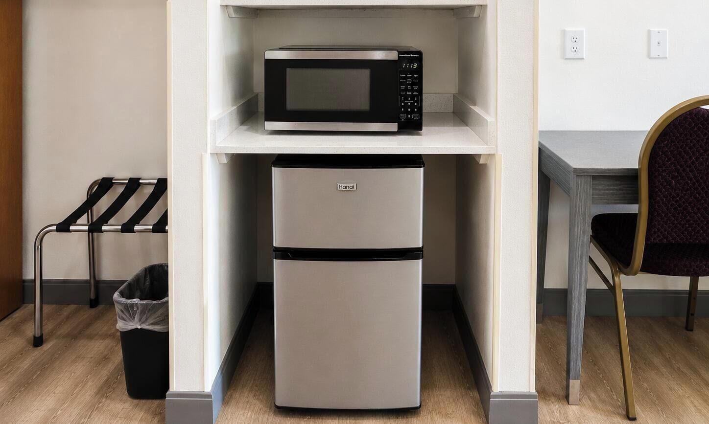 A compact kitchenette with a microwave and a refrigerator, a chair, and a trash bin.