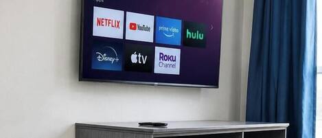 Una habitación moderna con una televisión de pantalla plana que muestra varias aplicaciones de streaming, un mueble de entretenimiento gris, un sillón tapizado morado y una ventana con cortinas.