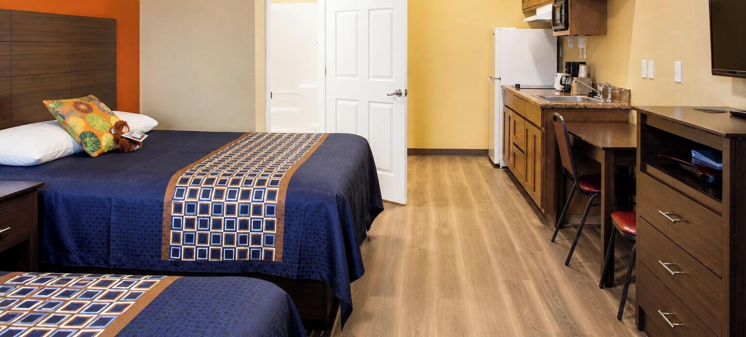 A hotel room with two beds, a desk, a chair, a television, and a kitchenette.