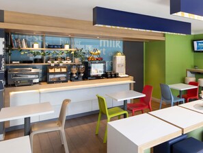 A breakfast bar with coffee machines, a fruit display, and a sign reading 'burger milk.' There are colorful chairs and tables.
