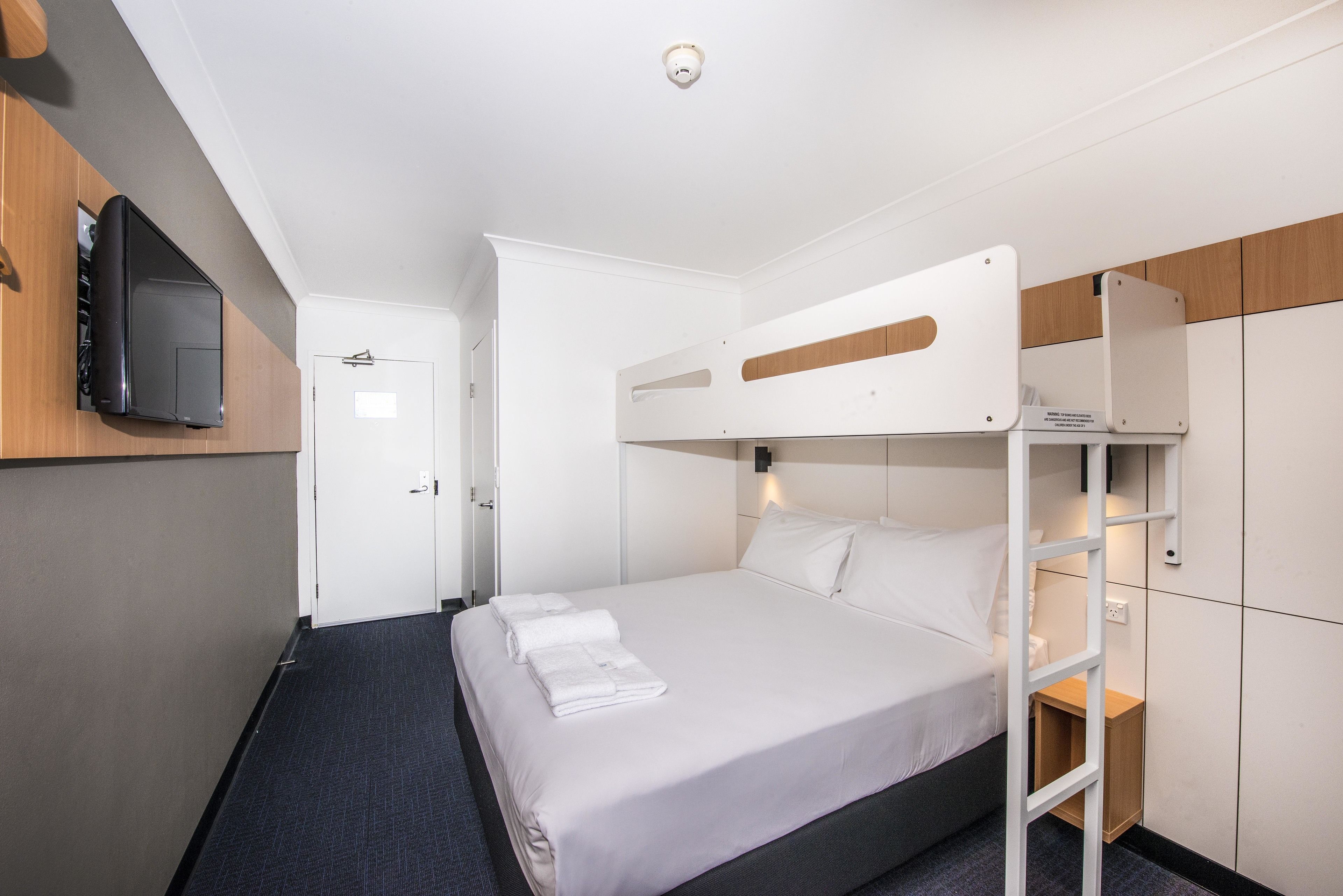 Habitación de hotel pequeña con una cama litera, un televisor de pantalla plana montado en la pared y un armario de madera.