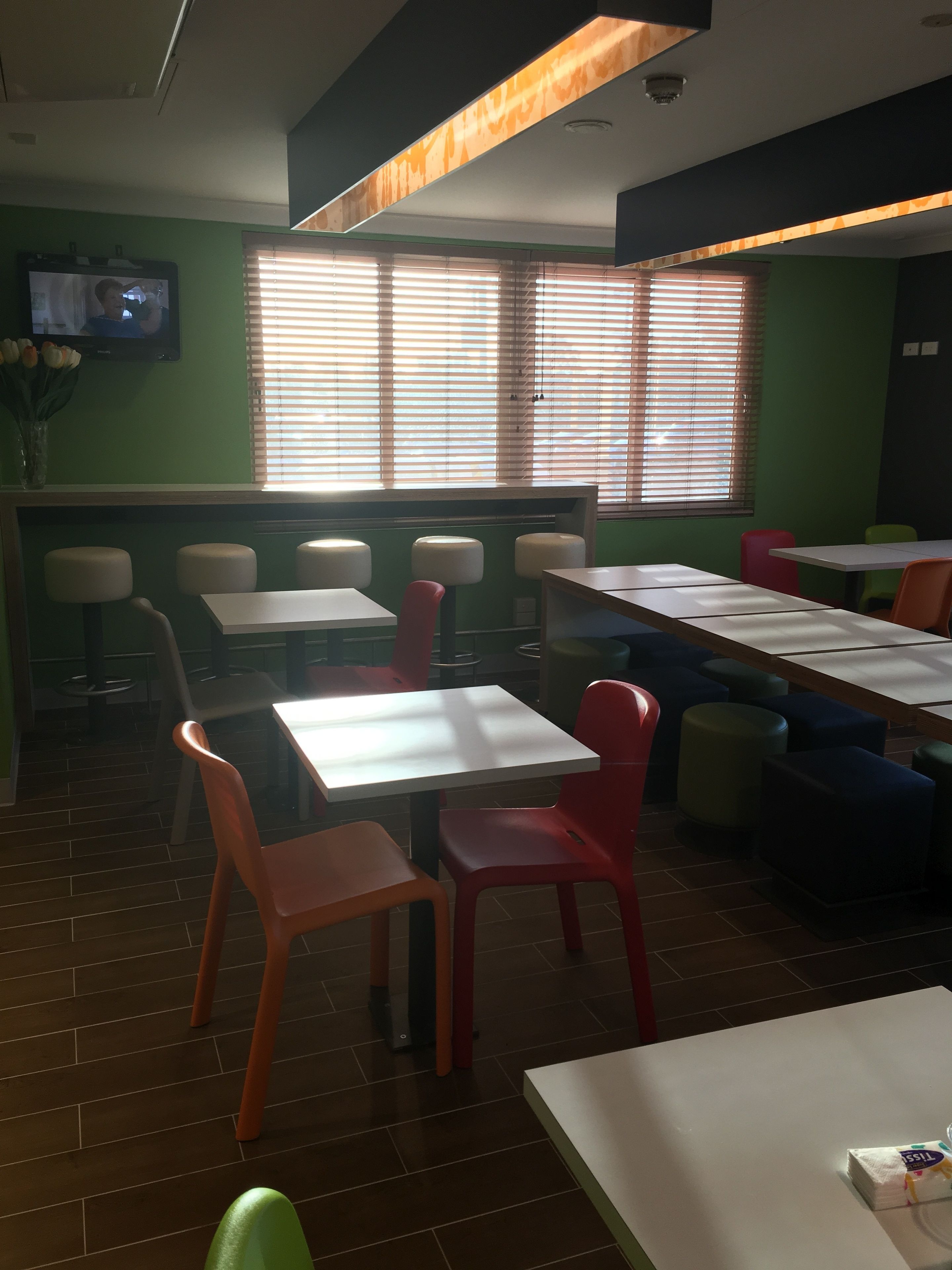 A restaurant interior with multiple tables and chairs, a television mounted on the wall, and a window with blinds.