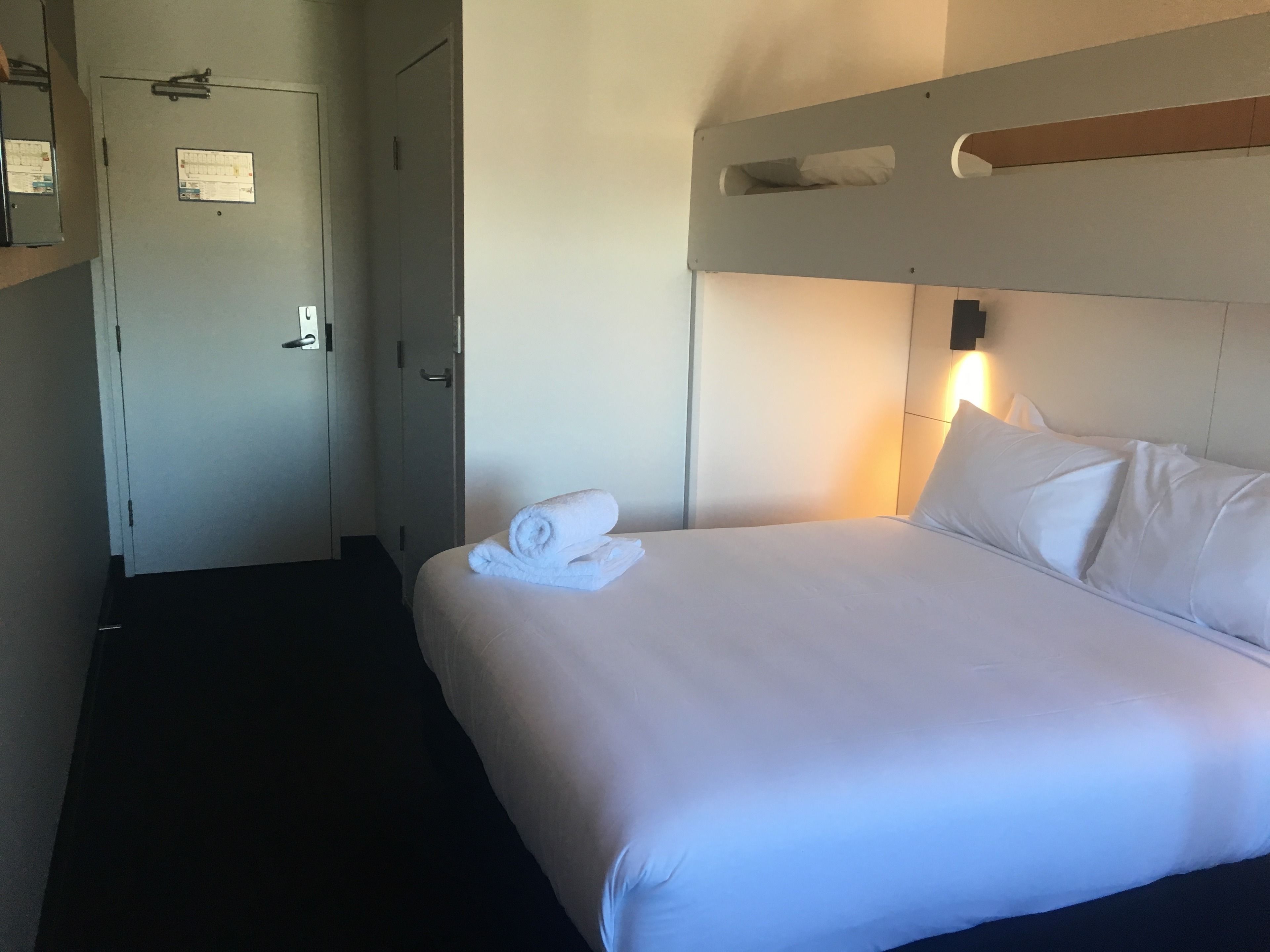 A hotel room with a neatly made bed, a bedside table, and a wall-mounted light fixture.