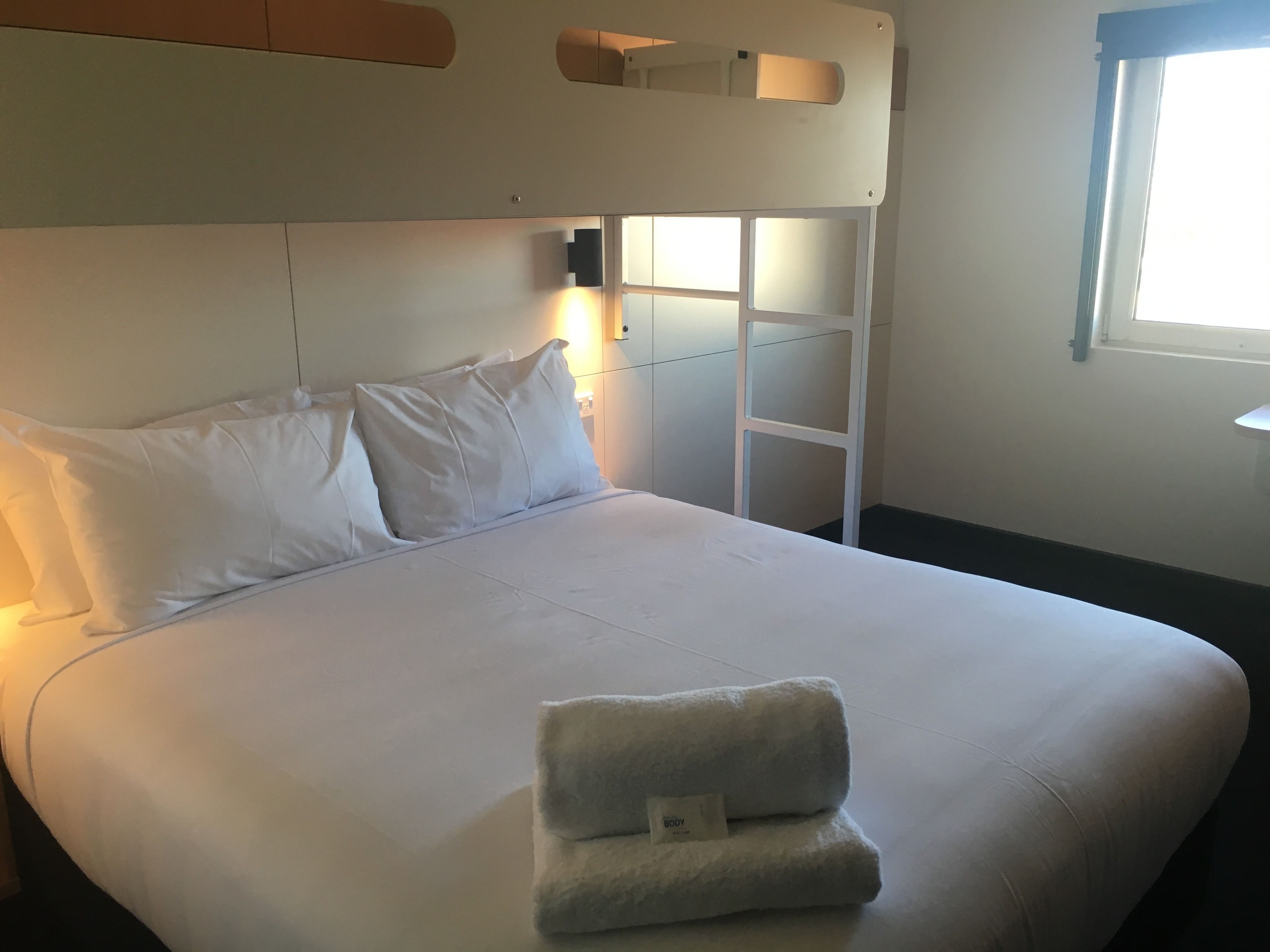 A neatly made bed with white linens and pillows, a folded towel on the bed, and a white headboard with a shelf.