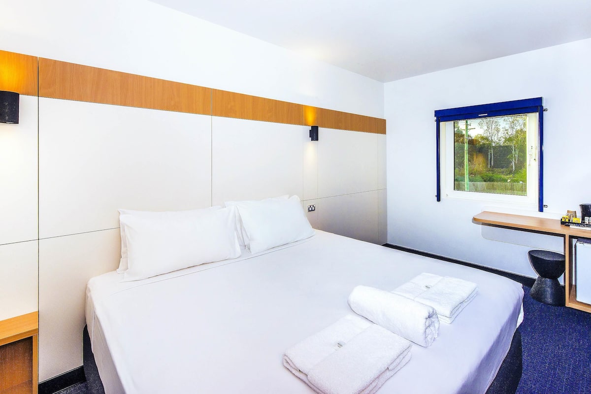 A hotel room with a large bed, a desk, and a window with a view of greenery.