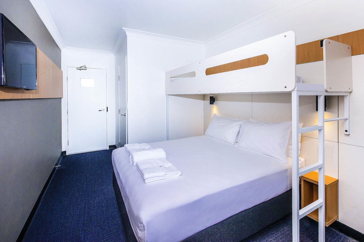 A bunk bed room with a television mounted on the wall, a door, and a small wooden side table.